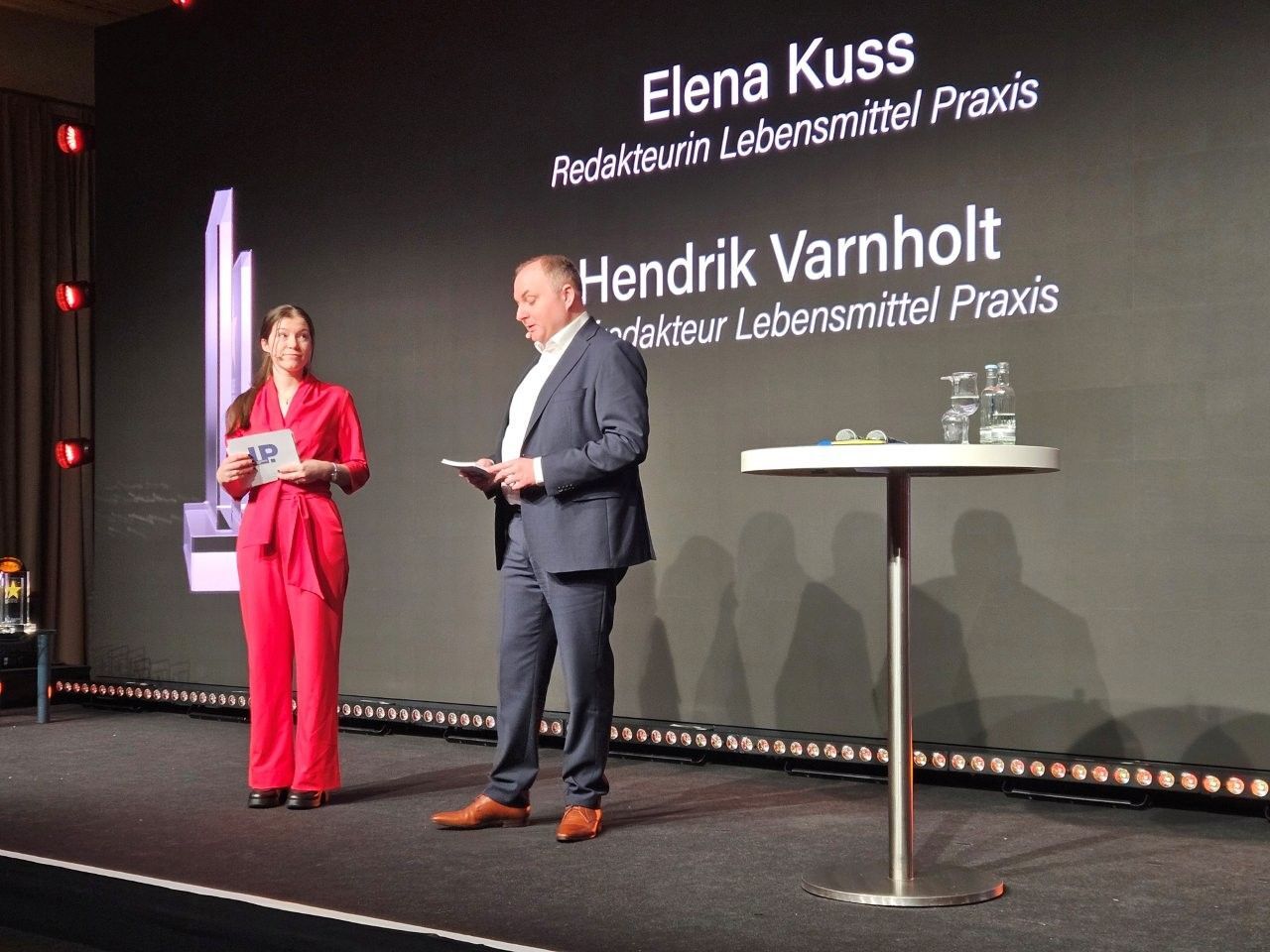 Zwei Personen auf der Bühne: eine Frau in Rot, ein Mann im Anzug, die bei einer Veranstaltung sprechen, Namen und Titel werden eingeblendet.