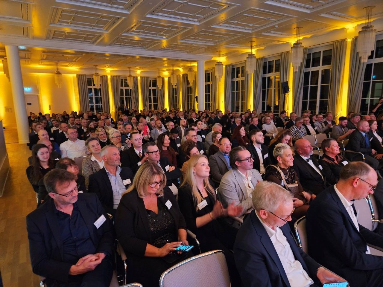 Publikum sitzt in einem großen Saal und lauscht; warmes Licht und hohe Decke.