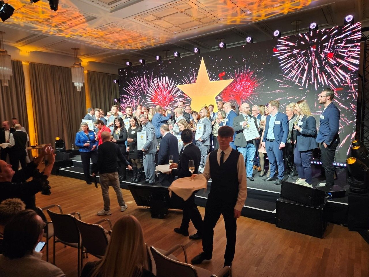 Bei einer Veranstaltung stehen Menschen auf einer Bühne vor einer Leinwand, auf der ein Stern und ein Feuerwerk zu sehen sind. Die Zuschauer stehen und schauen zu.