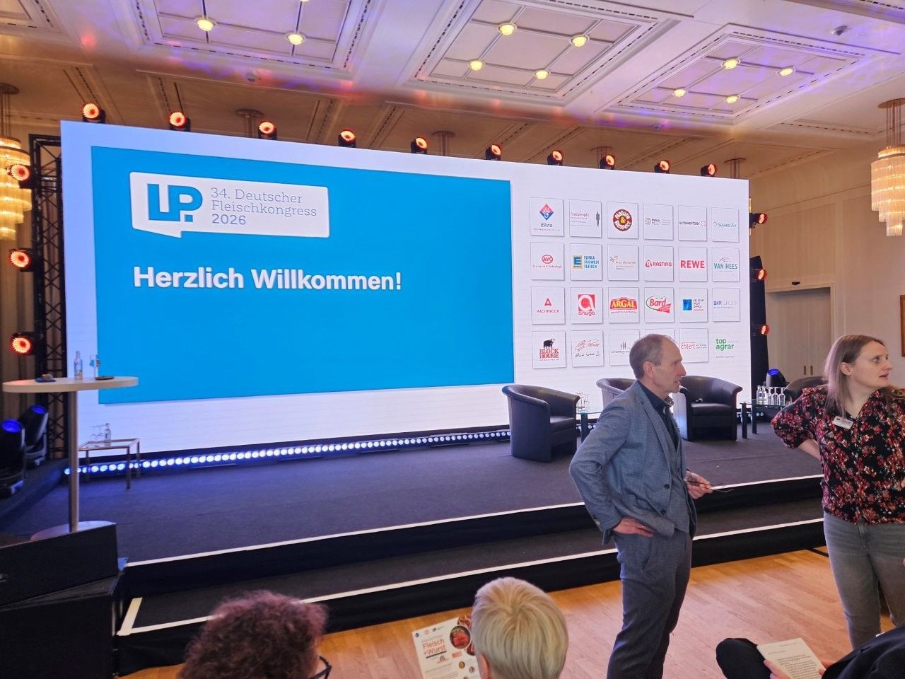 Konferenzbühne mit der Aufschrift „Herzlich Willkommen!“ auf einer Leinwand. Zwei Personen stehen daneben.
