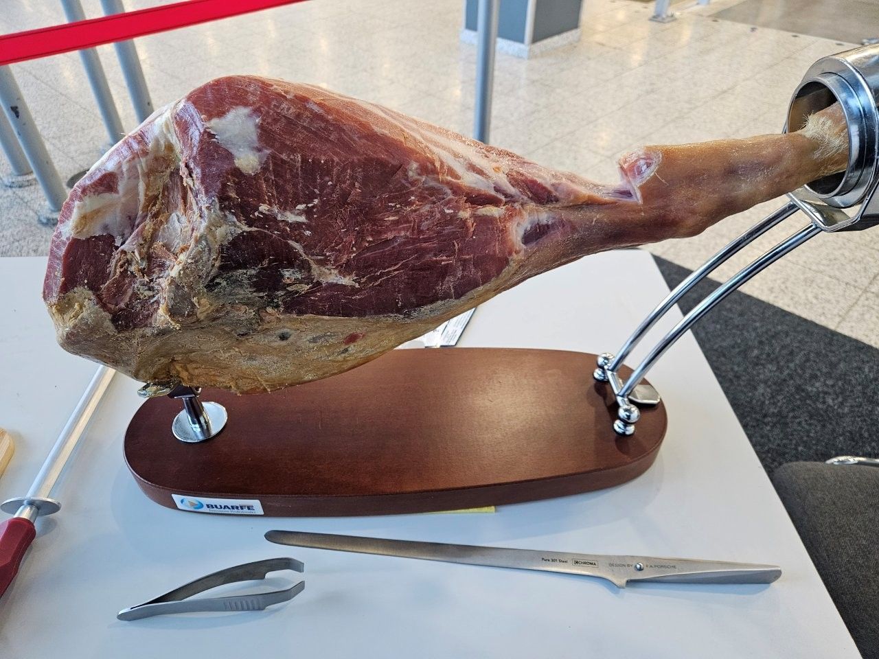 Gepökelter Schinkenschenkel auf einem Holzständer, dazu ein Tranchiermesser, eine Pinzette und ein Wetzstahl.