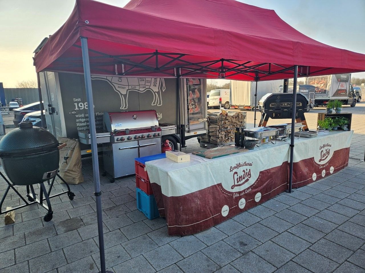 Ein rot überdachter Imbissstand mit Grill, Big Green Egg Smoker und Theke. Das Logo von „Lindi’s“ prangt auf der Theke.