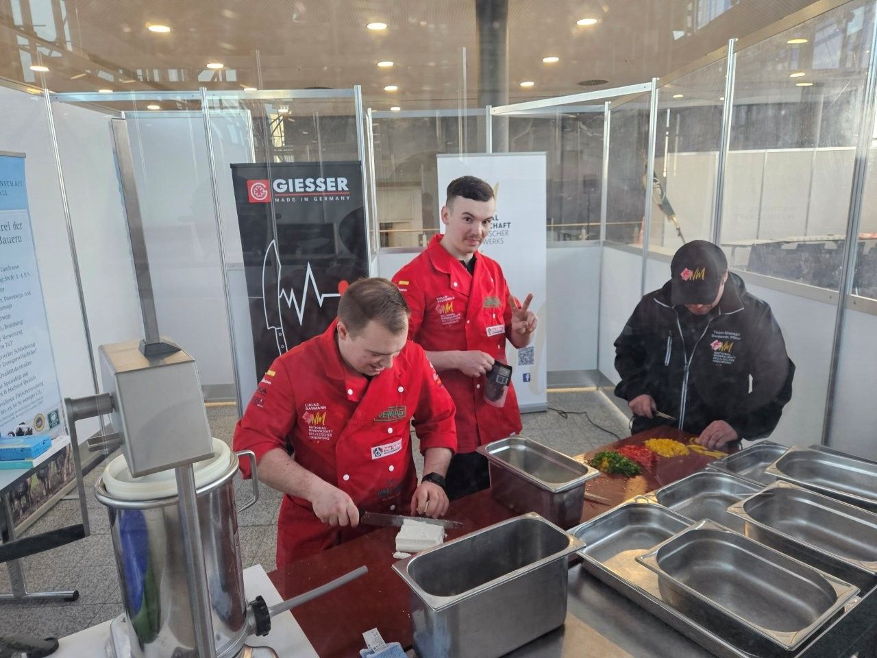 Drei Personen in roten Kochjacken bereiten an einer Theke Speisen zu, eine davon zeigt den Daumen nach oben.