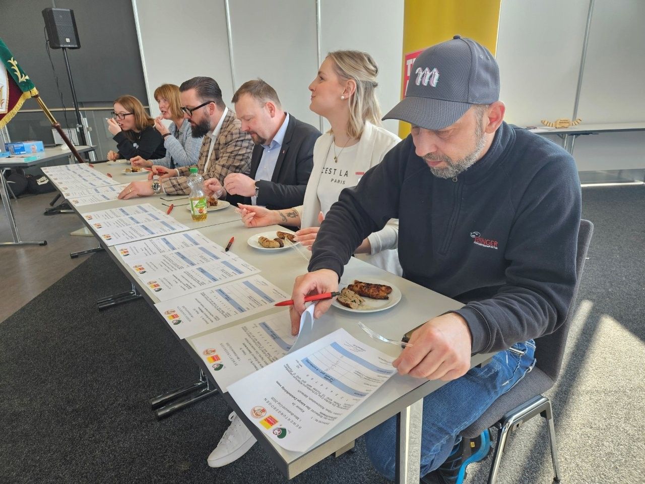 Menschen sitzen an einem langen Tisch und lesen Papiere durch; neben ihnen stehen Teller mit Essen. Eine Person trägt eine Baseballkappe und untersucht ein Dokument.