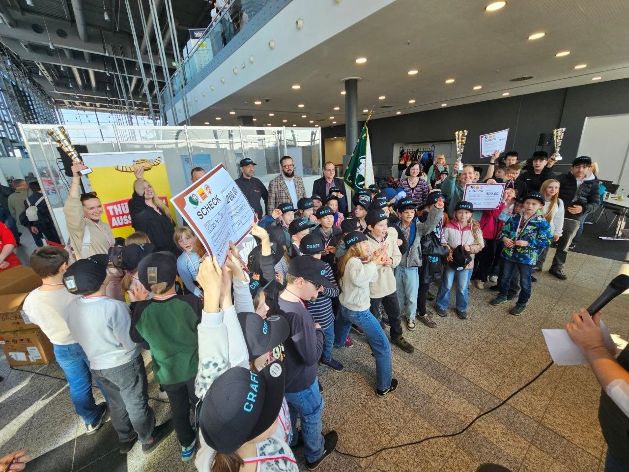 Kinder feiern mit einer Urkunde in einem modernen Gebäude, umgeben von Menschen und Trophäen.