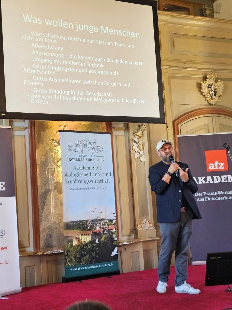 Redner am Rednerpult in einem prunkvollen Saal, Präsentation neben einer projizierten Folie, im Vordergrund sitzendes Publikum.