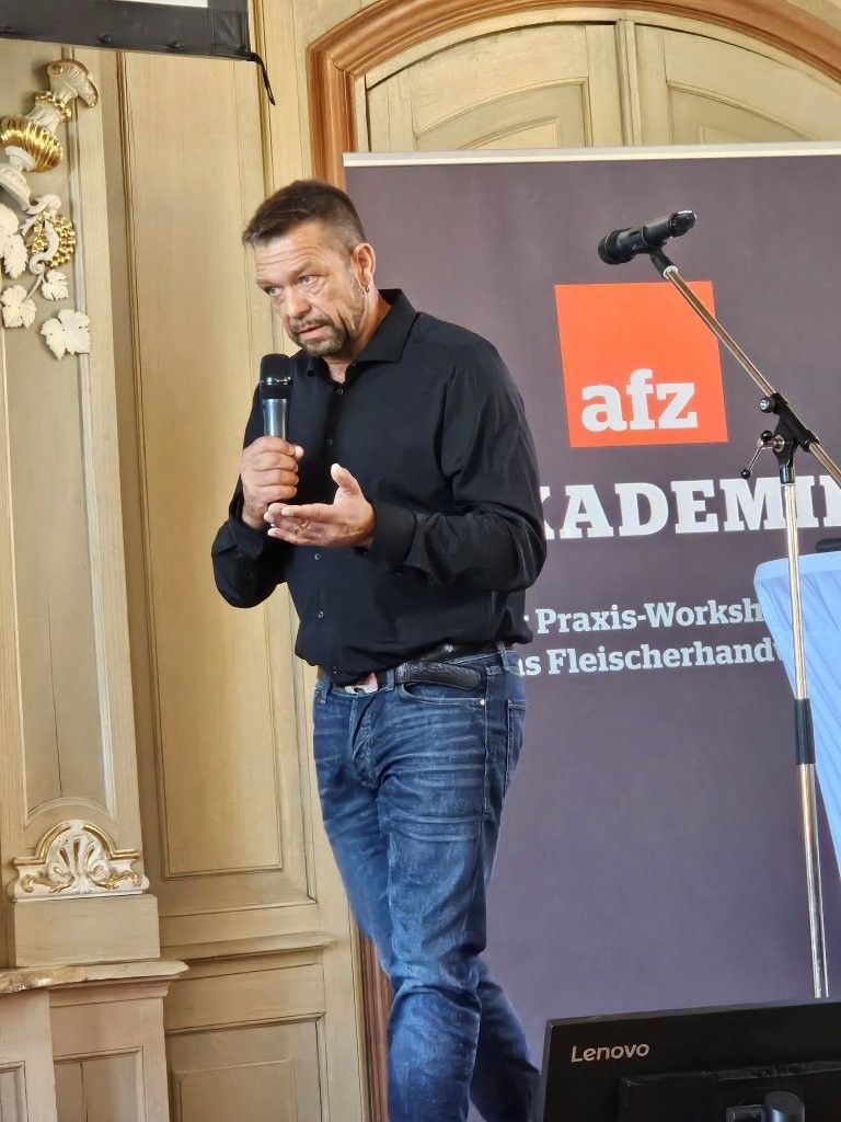 Ein Mann spricht neben einem Banner der AFZ Academy auf einer Bühne in ein Mikrofon.