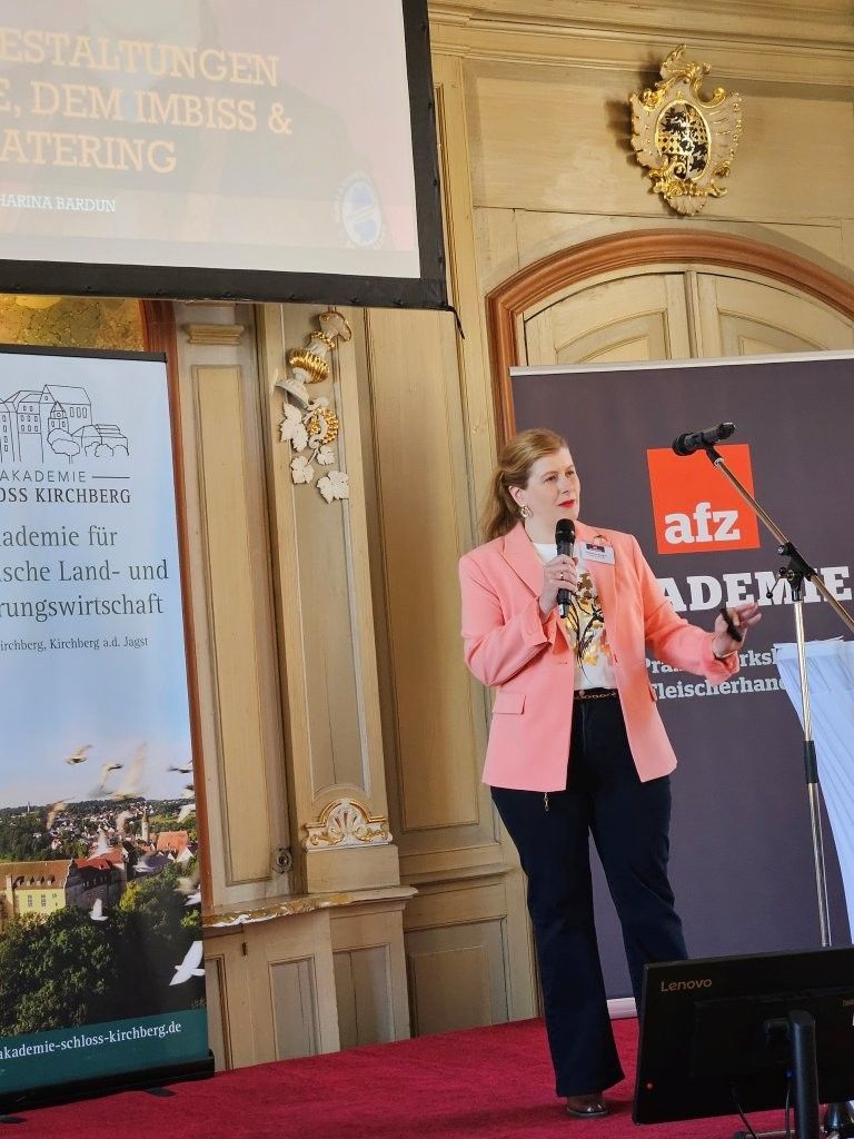 Frau im rosa Blazer spricht auf einem Podium auf einer mit rotem Teppich ausgelegten Bühne in einem Innenraum.