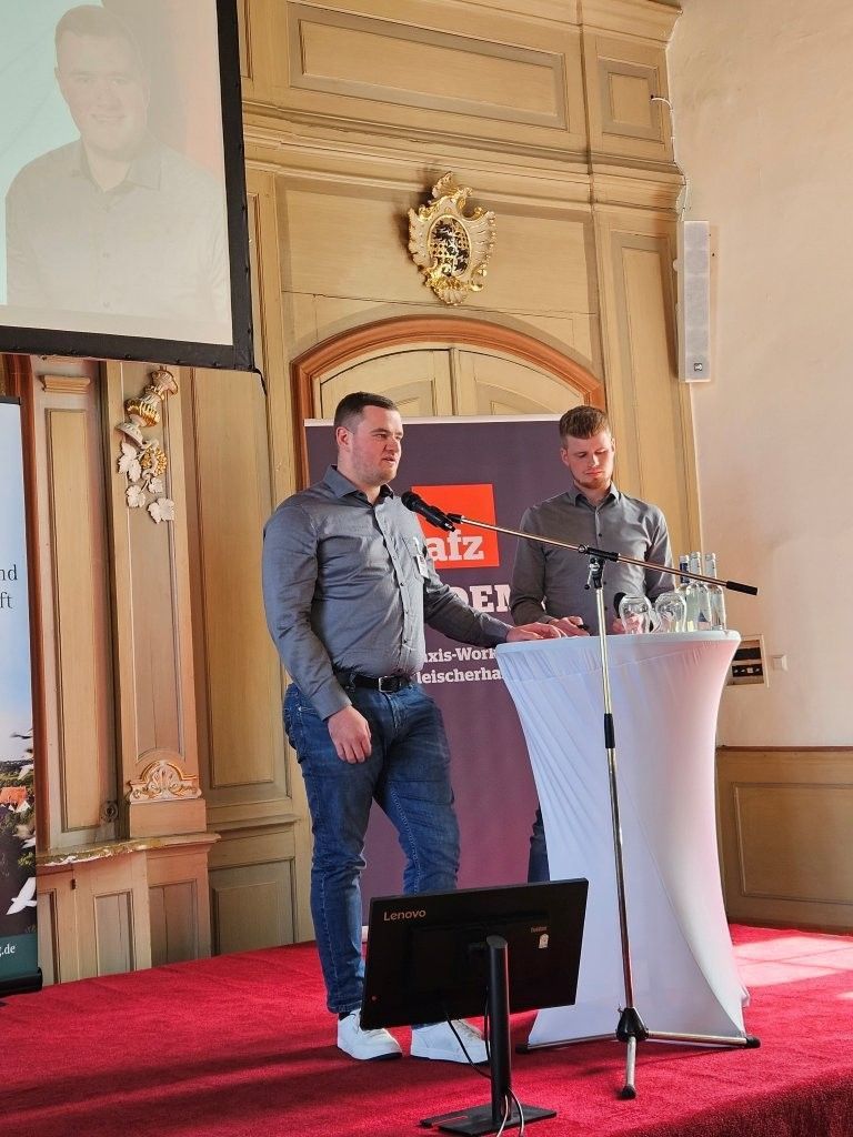 Zwei Männer sprechen an einem Rednerpult auf einer mit rotem Teppich ausgelegten Bühne in einem prunkvollen Saal; hinter ihnen befindet sich eine Leinwand.