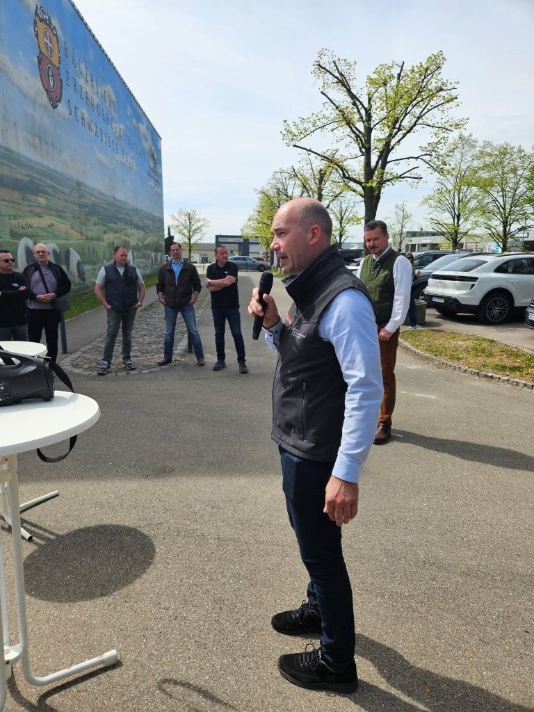 Ein Mann in schwarzer Weste spricht im Freien zu einer kleinen Gruppe in der Nähe von geparkten Autos und einer Wandmalerei.