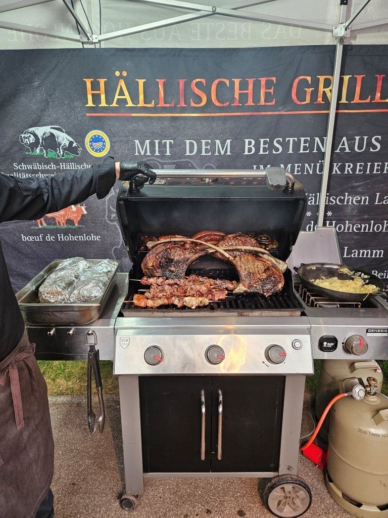 Grillstand im Freien mit schwarzem Smoker und aufgeschnittenem Bratenfleisch vor einem Banner mit der Aufschrift „Hällischer Grill“.