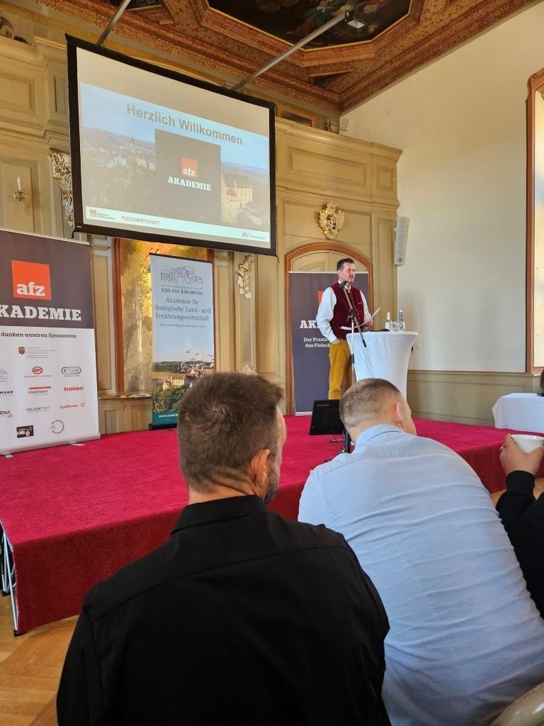 Redner am Rednerpult in einem prunkvollen Saal, spricht zu einem sitzenden Publikum, während die Präsentation auf eine Leinwand projiziert wird.