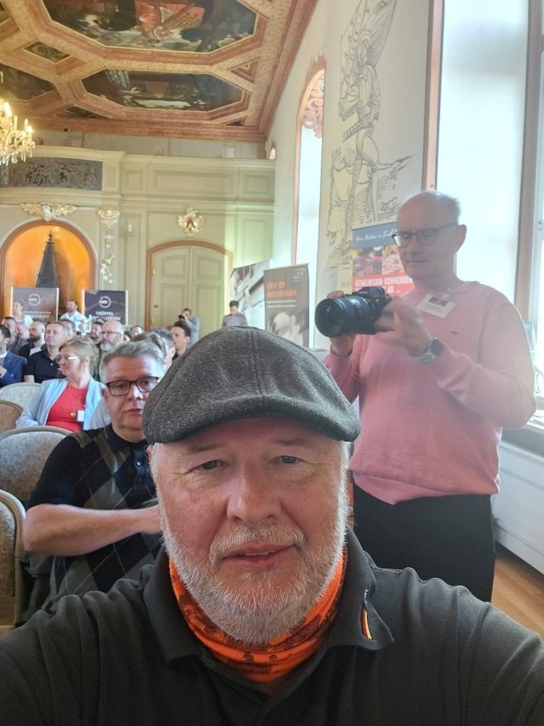Ein Mann mit Mütze und orangefarbenem Schlauchschal sitzt in einer reich verzierten Halle; hinter ihm steht ein Fotograf.