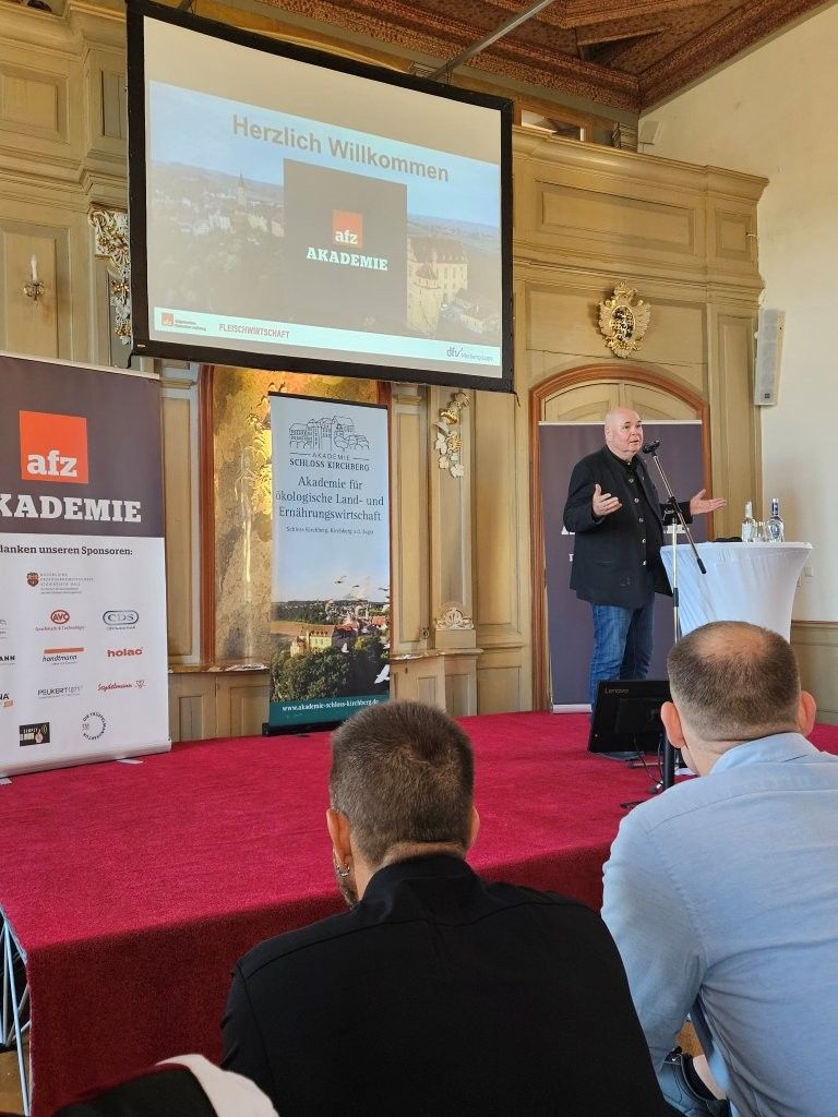 Ein Redner präsentiert seinen Vortrag an einem Konferenzpult in einem prunkvollen Saal; eine Folie wird projiziert, und das Publikum sitzt davor.