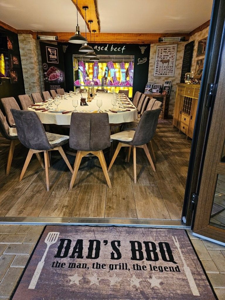 Esszimmer mit rundem Tisch und Stühlen, das sich zu einem Grillplatz im Garten mit einer „DAD'S BBQ“-Grillmatte öffnet.