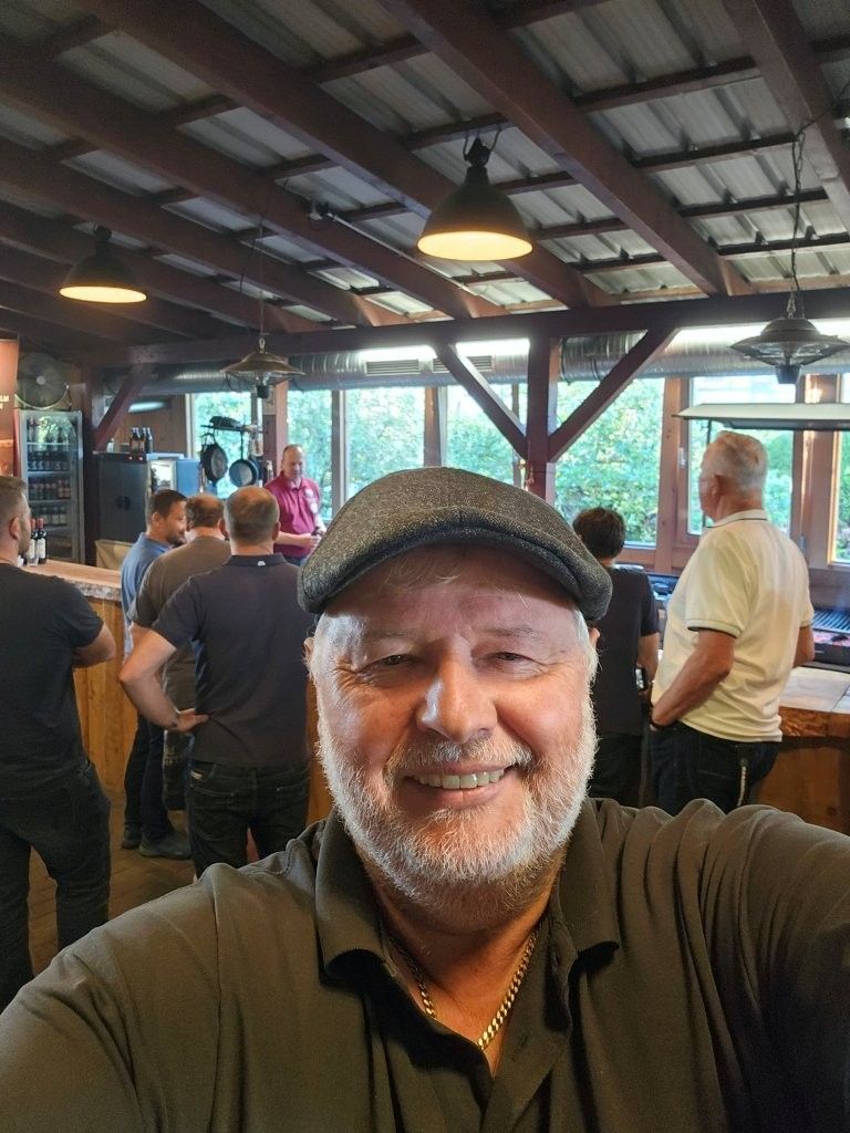 Ein lächelnder Mann macht ein Selfie in einer belebten, überdachten Bar im Freien, im Hintergrund Menschen und warmes Licht.