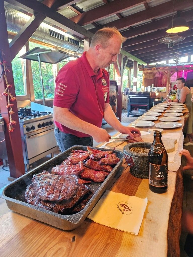 Ein Koch tranchiert Grillfleisch an einem Buffet mit Tellern und Tabletts unter einer rustikalen, überdachten Terrasse.