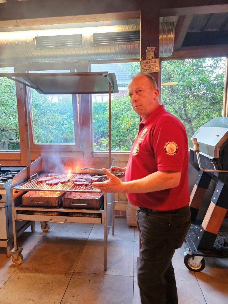 Ein Mann in einem roten Hemd grillt Essen neben einem angezündeten Grill in einem überdachten Außenbereich.
