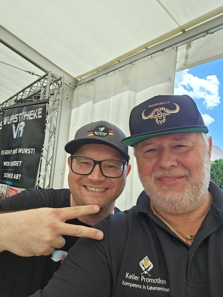 Zwei lächelnde Männer mit Kappen machen ein Selfie an einem Stand im Freien.