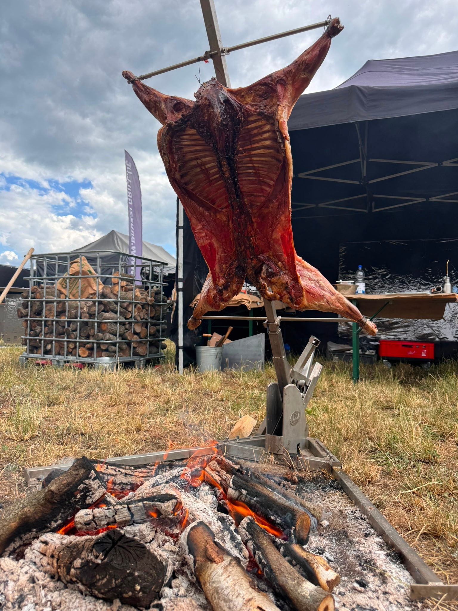 Ein ganzes Tier wird im Freien am Spieß über offenem Feuer gebraten, im Hintergrund ist ein Bühnenzelt zu sehen.