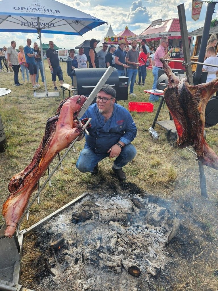 Ein Mann kauert an einem offenen Feuer und hält ein gebratenes ganzes Schwein bei einer Feier im Freien mit Menschen und Zelten.