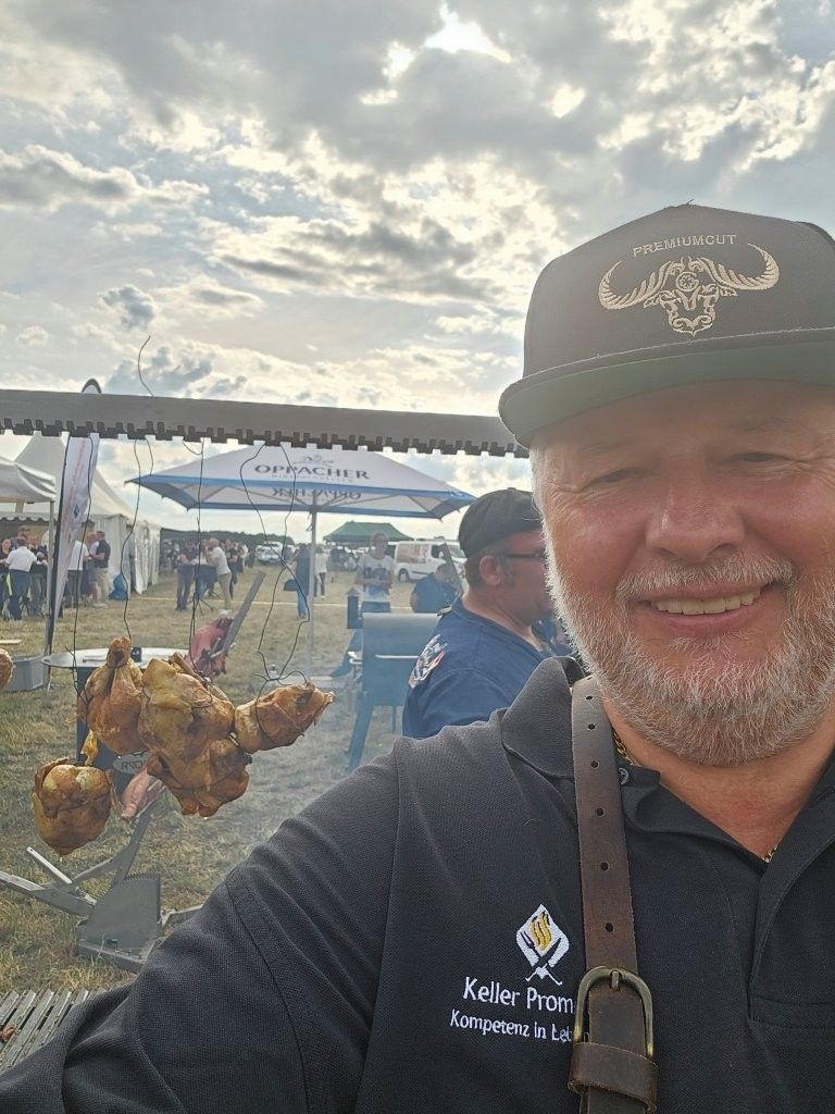 Ein lächelnder Mann mit schwarzer Kappe hält gegrilltes Hähnchen auf einem Freiluft-Food-Festival unter bewölktem Himmel.