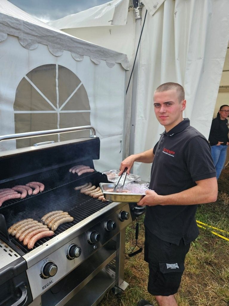 Ein Mann grillt Würstchen an einem Freiluftbuffet neben einem weißen Veranstaltungszelt.