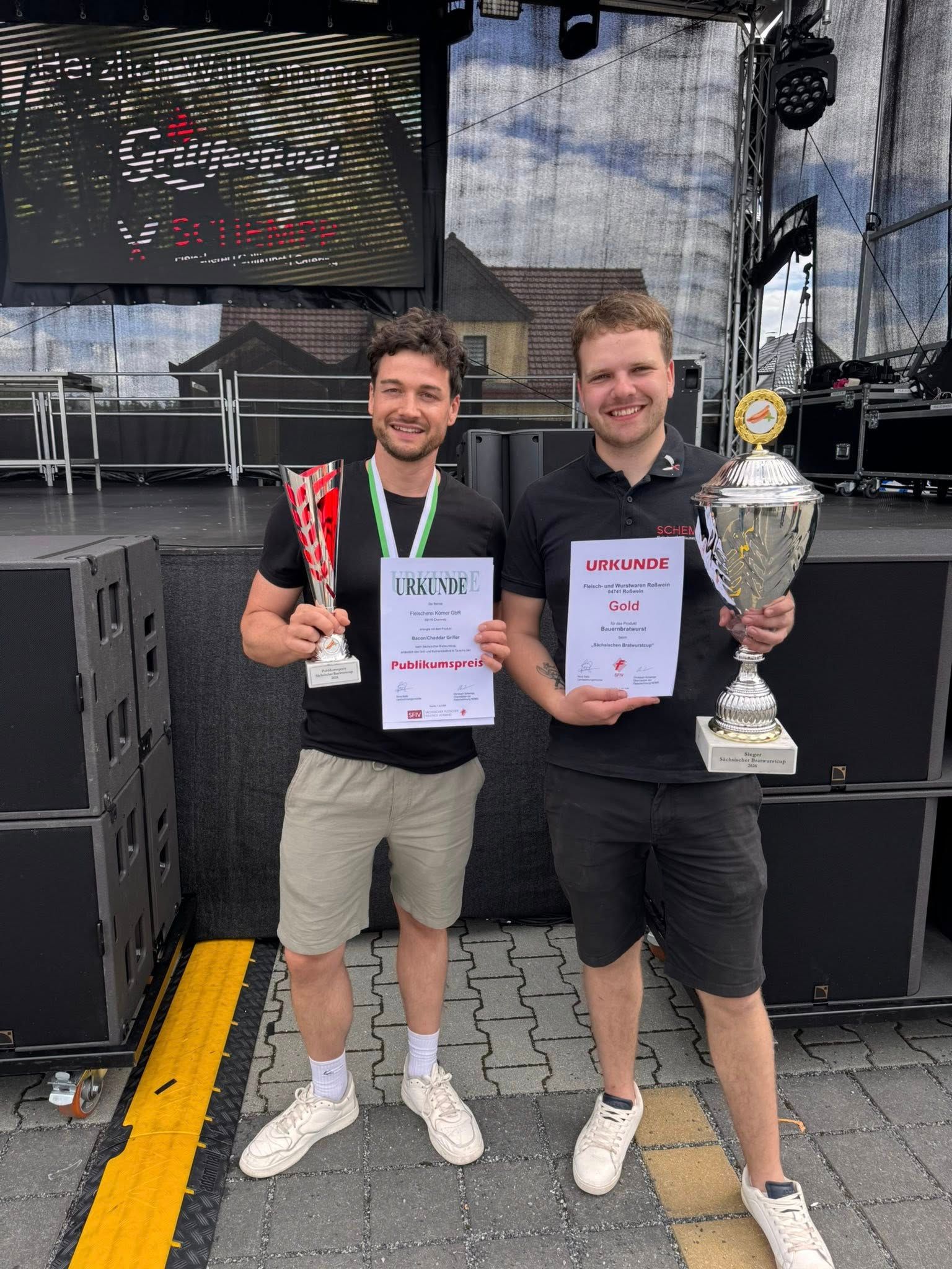 Zwei Männer, die nach einem Wettbewerb Urkunden und einen Pokal auf einer Bühne halten und lächeln.