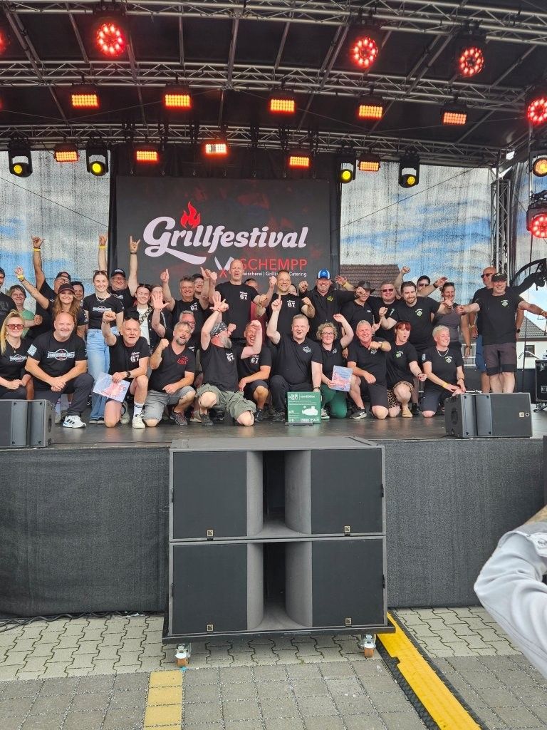 Eine Gruppe von Menschen posiert auf einer Freilichtbühne unter rotem Licht auf einem Banner des Grillfestivals.