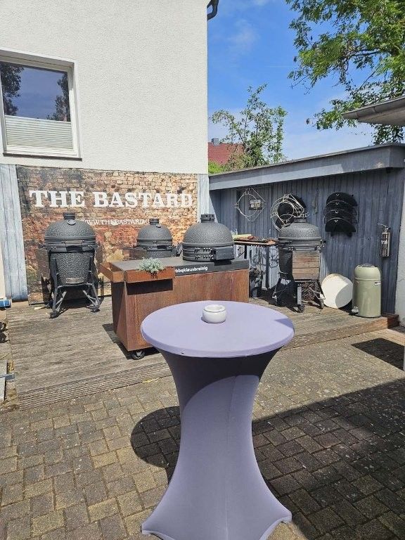 Terrasse im Freien mit einem lila Cocktailtisch, Grillmöglichkeiten und einem Backsteinmauerschild mit der Aufschrift „DER BASTARD“
