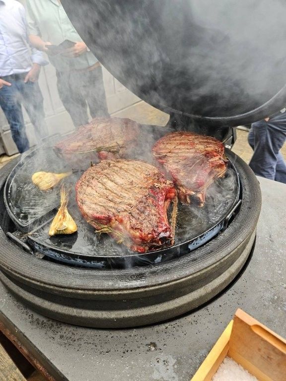 Steaks und Maiskolben werden auf einem runden Holzkohlegrill gegrillt, Rauch steigt auf – draußen beim Barbecue.