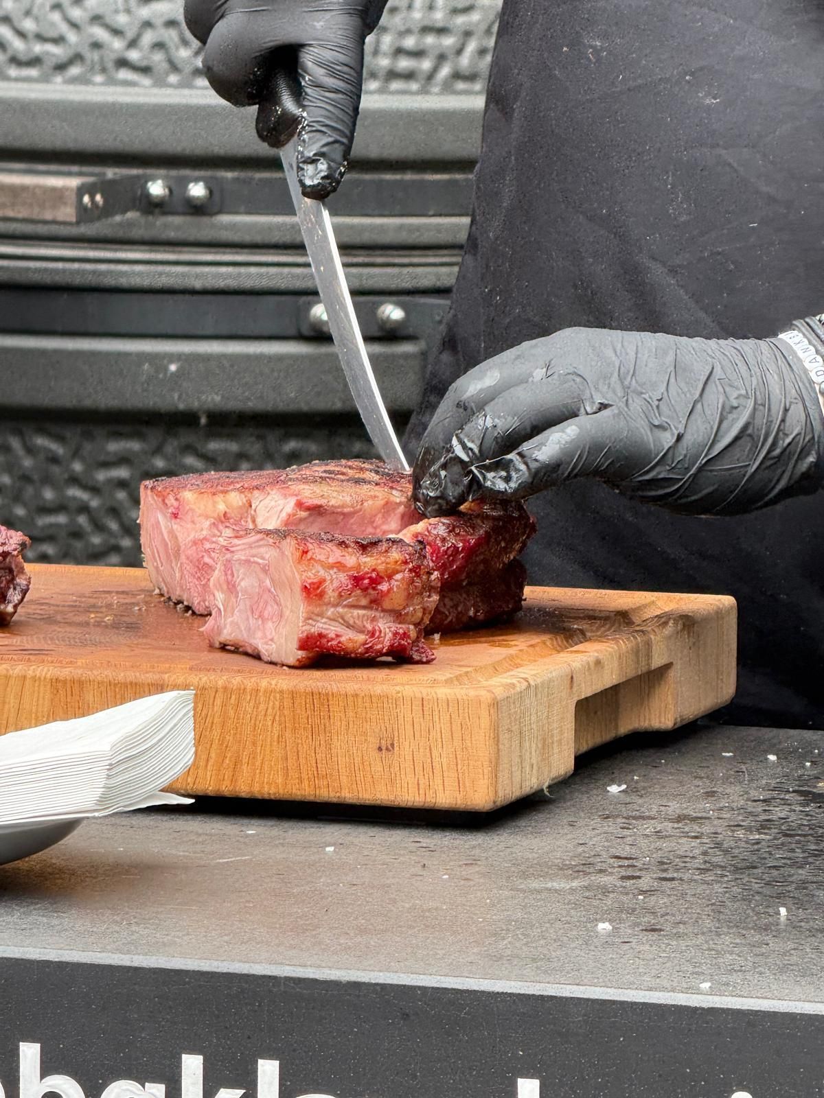 Behandschuhte Hände schneiden draußen ein Stück gegrilltes Fleisch auf einem Holzbrett.