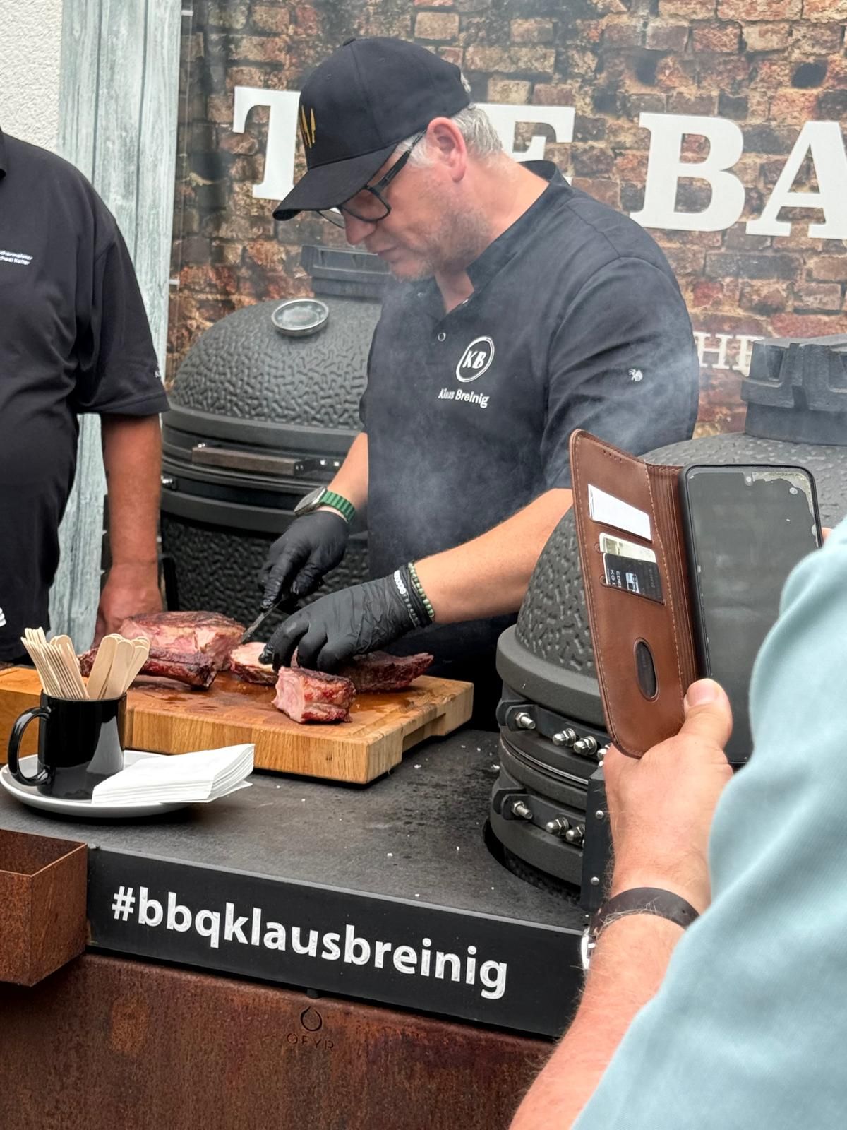Mann mit schwarzer Mütze und Handschuhen schneidet Fleisch an einer Grillstation; davor ist ein Hashtag-Schild zu sehen.