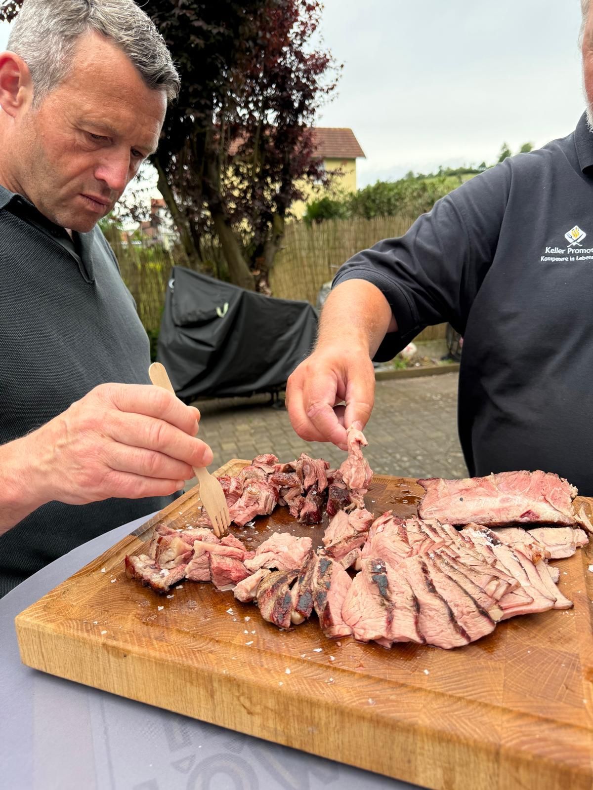 Zwei Personen schneiden im Freien Scheiben von Bratenfleisch auf einem Holzbrett.