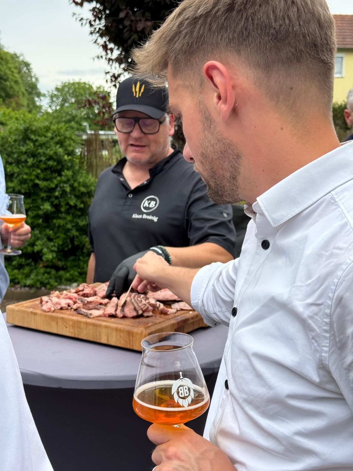 Zwei Männer beim Grillen im Freien, sie schneiden Fleisch und halten Getränke in der Nähe eines Holztisches.