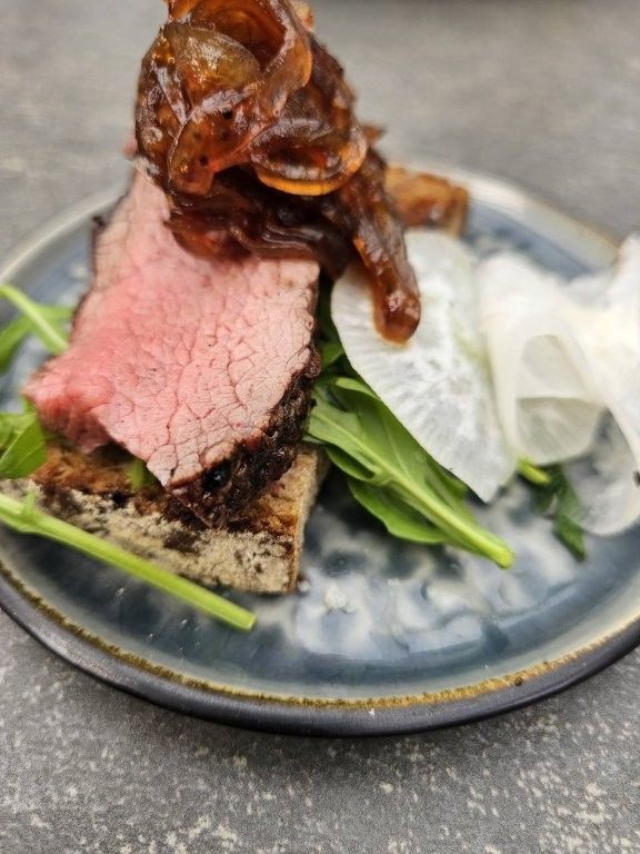 Eine Scheibe Roastbeef mit karamellisierten Zwiebeln auf Salat, daneben eingelegte Zwiebeln auf einem blaugrauen Teller.