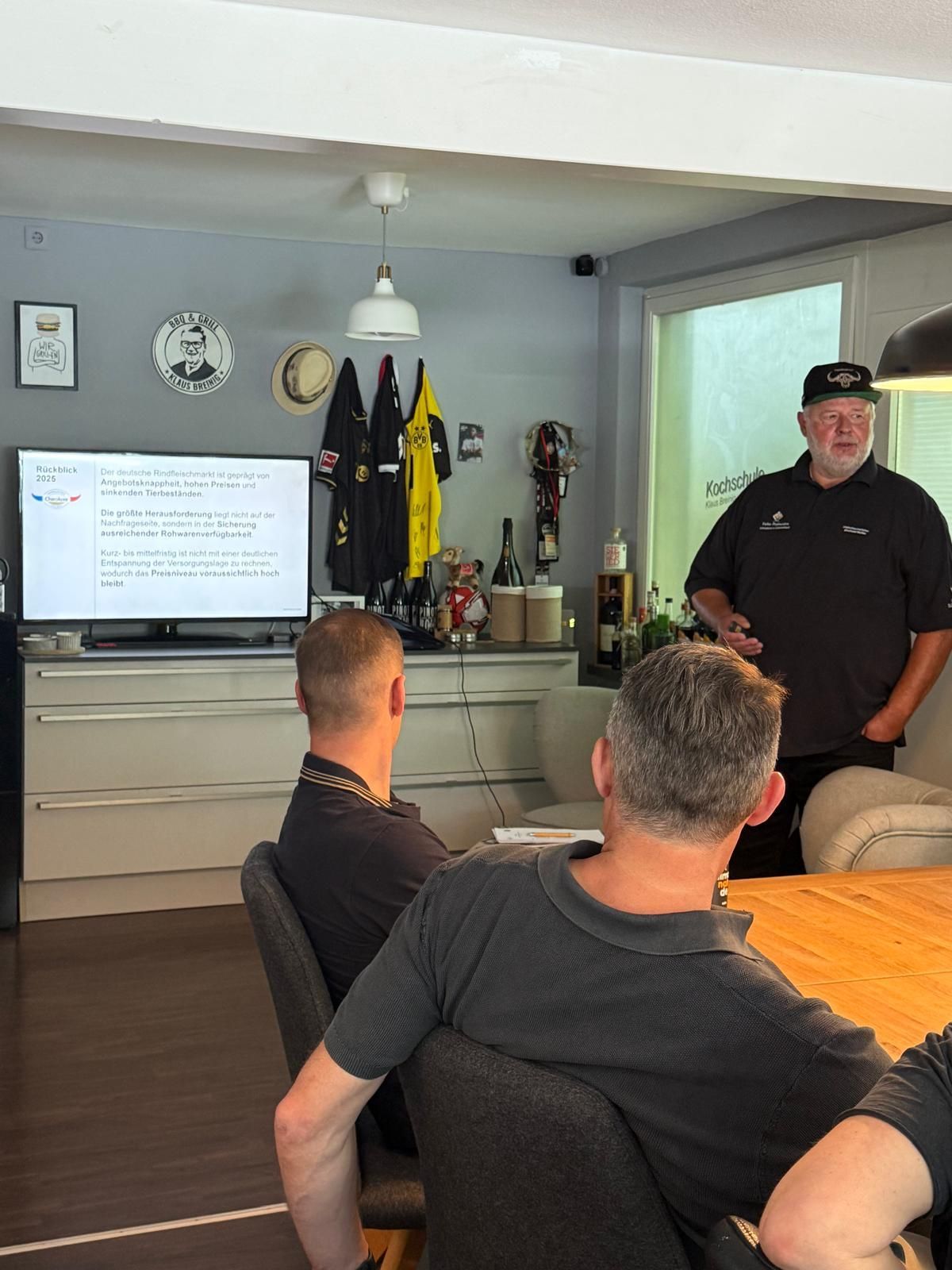Ein Mann spricht während einer Präsentation in einem Wohnzimmer; zwei Männer sitzen daneben, und in der Nähe wird eine Fernsehfolie gezeigt.