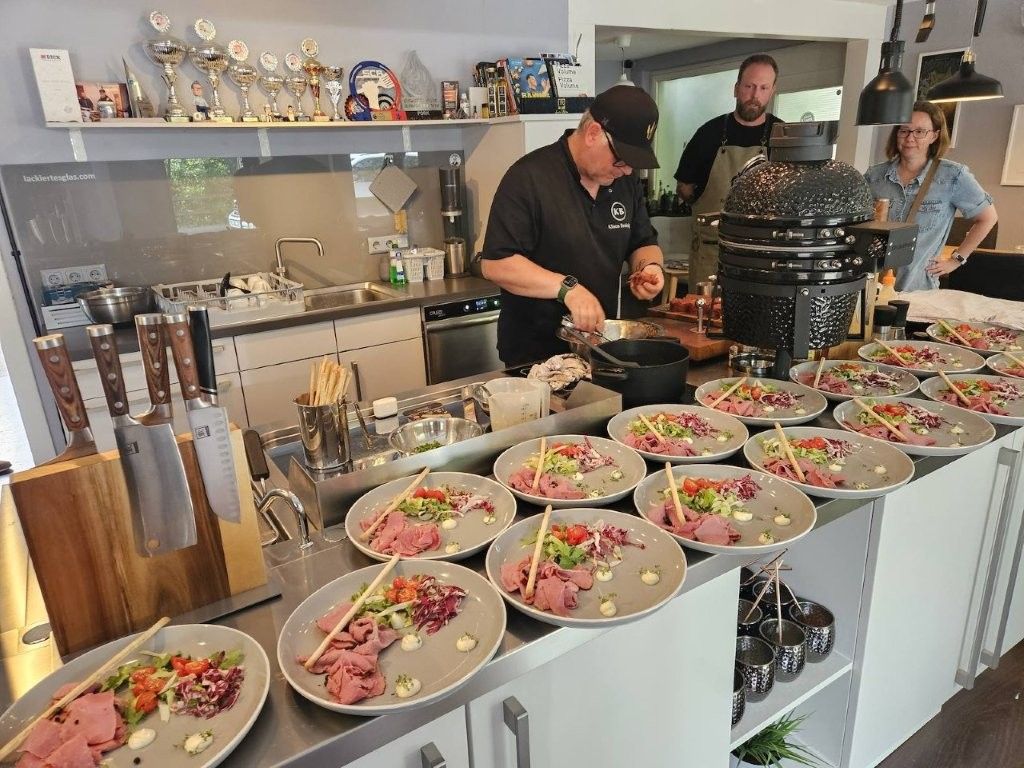 Buffet mit angerichteten Salaten und zwei Personen, die in einer Großküche Essen zubereiten
