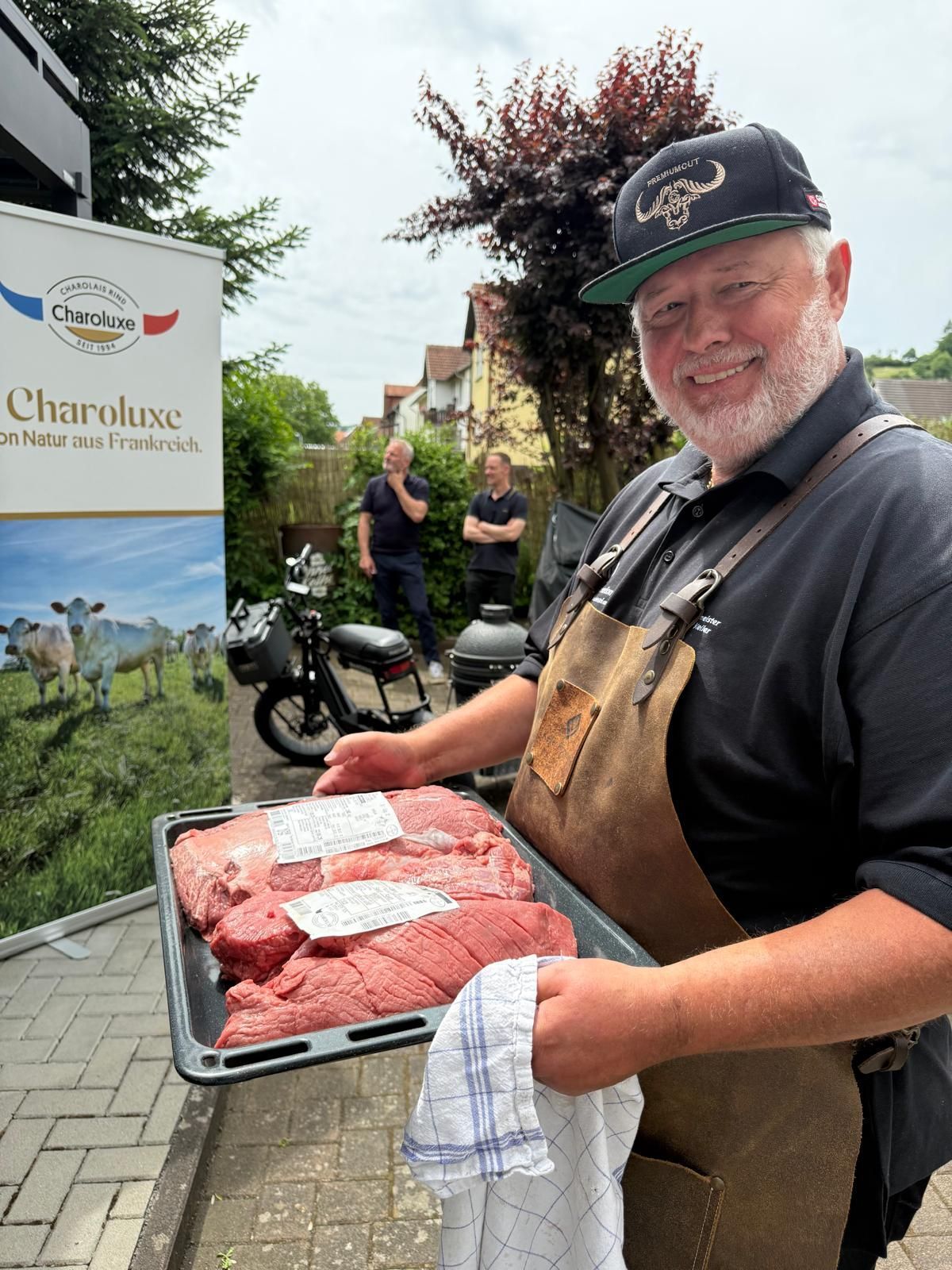 Ein Metzger lächelt, während er an einem Marktstand im Freien ein Tablett mit rohen Steaks hält.