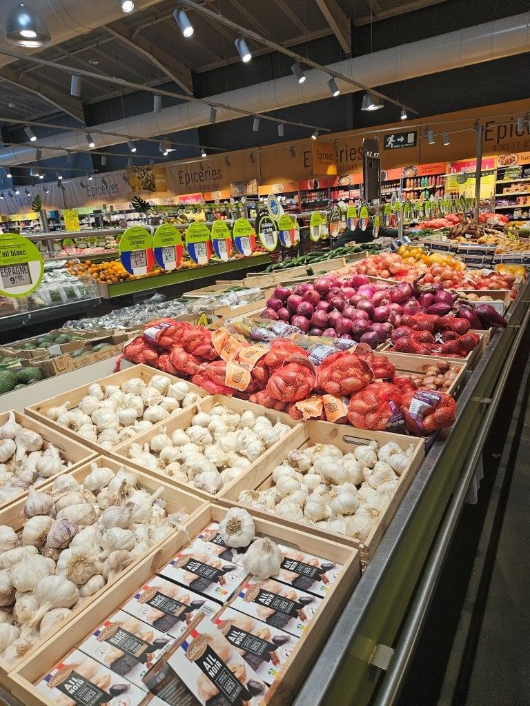 Die Obst- und Gemüseabteilung mit Kisten voller weißem Knoblauch, roten Zwiebeln und Kartoffeln unter hellen Ladenlampen