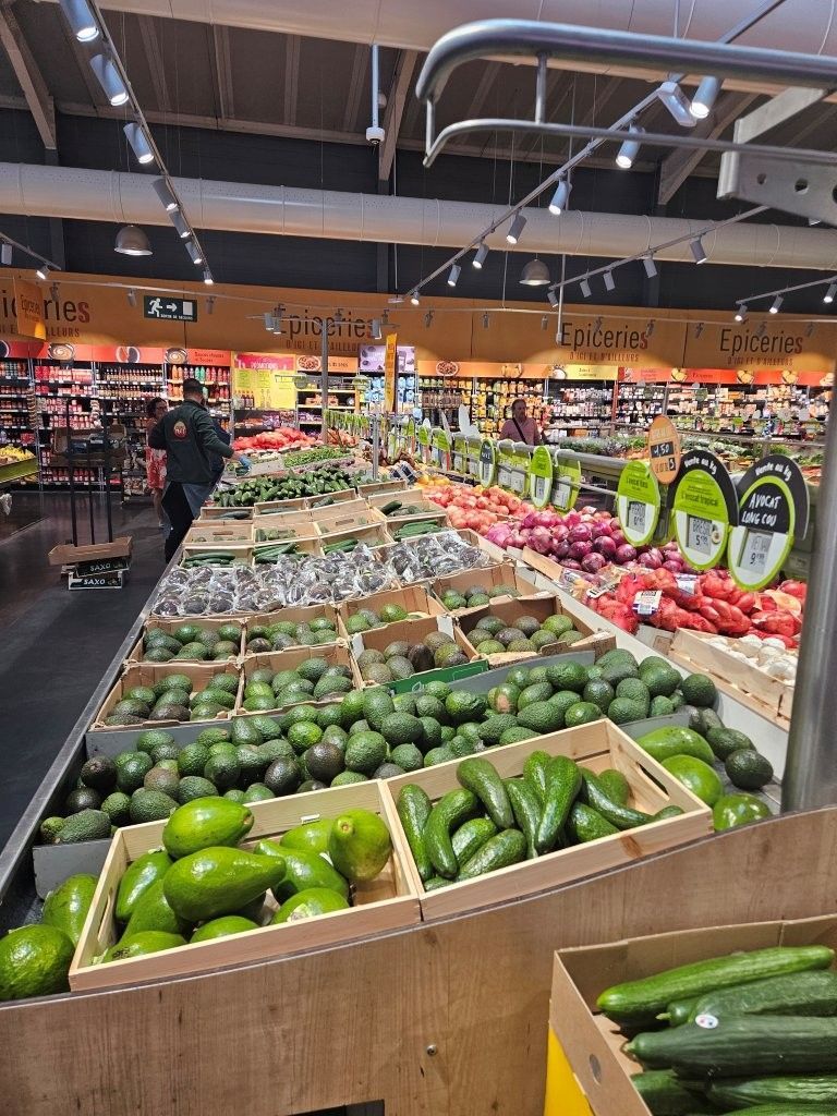 Die Obst- und Gemüseabteilung eines Supermarkts mit Avocados, Paprika und anderem frischen Gemüse, das auf Holztischen ausgestellt ist.