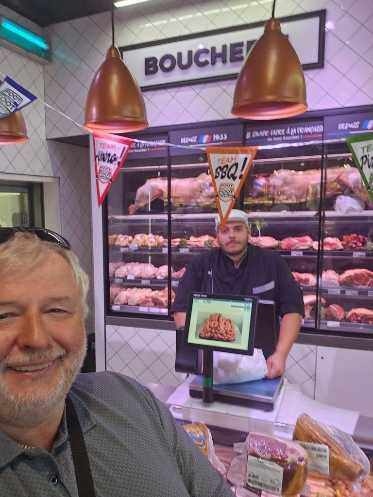 Theke einer Metzgerei mit einem Mann, der ein Tablett mit Fleischstücken hält, dahinter eine Vitrine und Hängelampen.