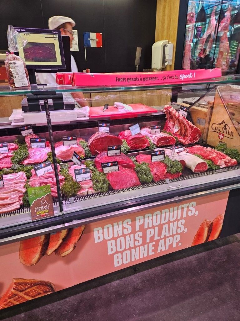 Fleischtheke mit verschiedenen Fleischsorten in einem Markt, mit einem Schild mit der Aufschrift „BONS PRODUITS. BONS PLANS. BONNE APP.“