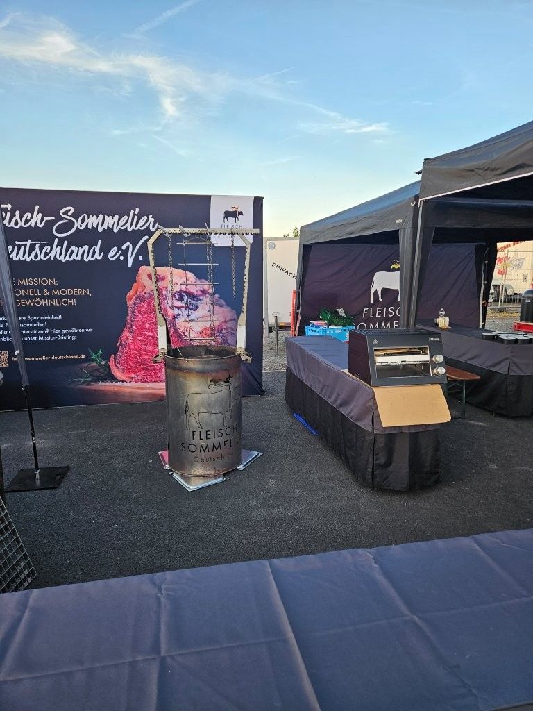 Messestand im Freien mit schwarzem Banner, pinkfarbener Grafik und Tischen unter Zelten auf einem gepflasterten Platz.