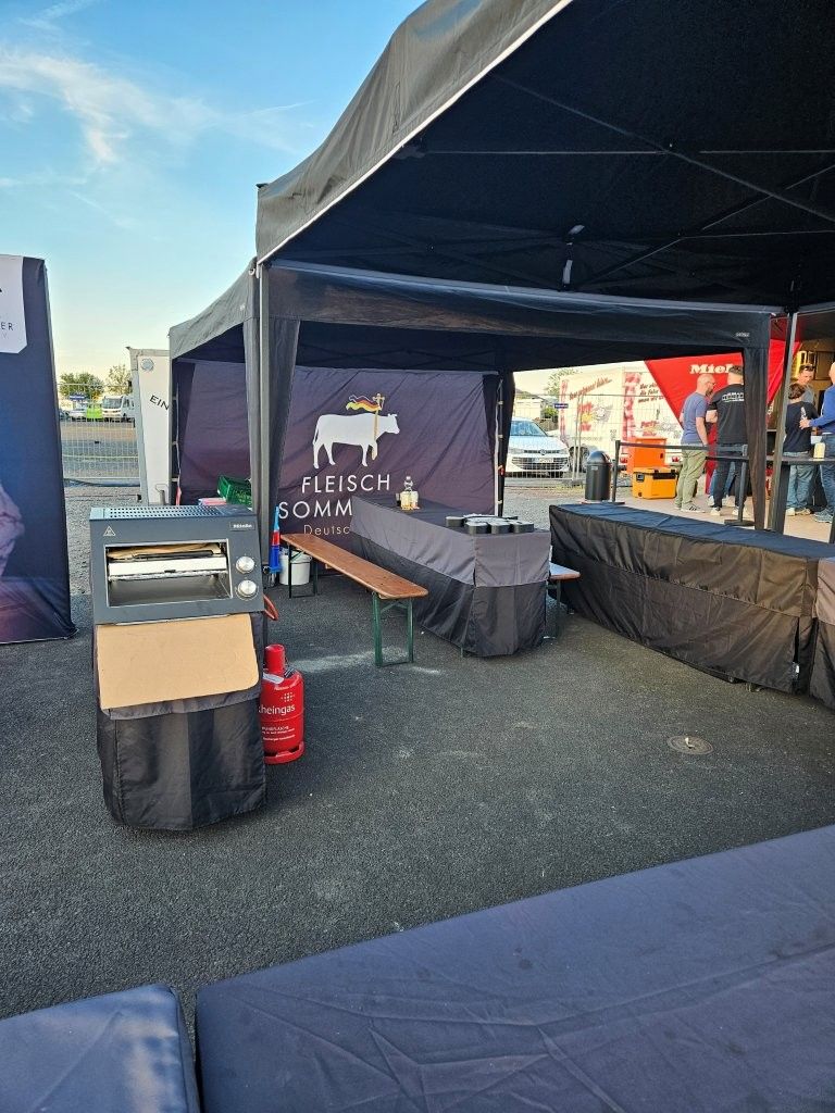 Im Freien aufgestellter Stand mit schwarzem Vordach, Tischen und rotem Feuerlöscher auf Kies auf einem Veranstaltungsgelände