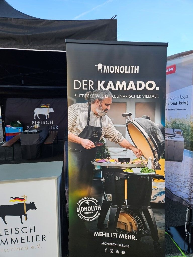 Monolith Kamado Grillstand mit Koch, der im Freien unter einem schwarzen Zelt kocht; Werbebanner und Logo sind sichtbar.
