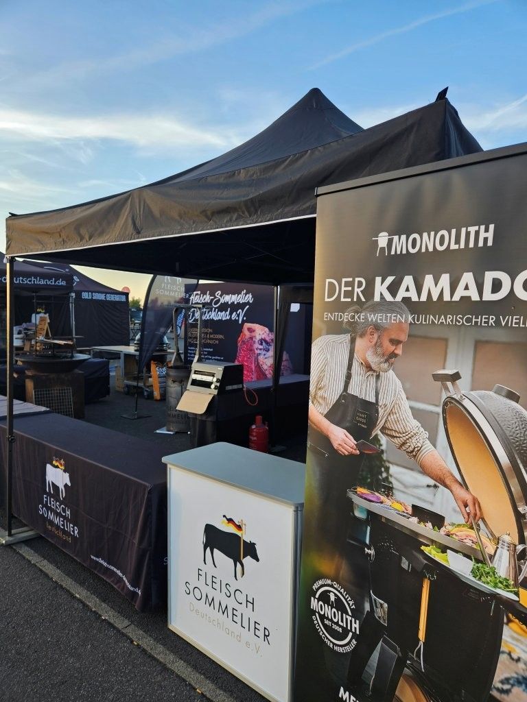 Kaffeestand im Freien mit schwarzen Pavillonzelten und dem Banner „Monolith Der Kamado“ bei einer Veranstaltung.