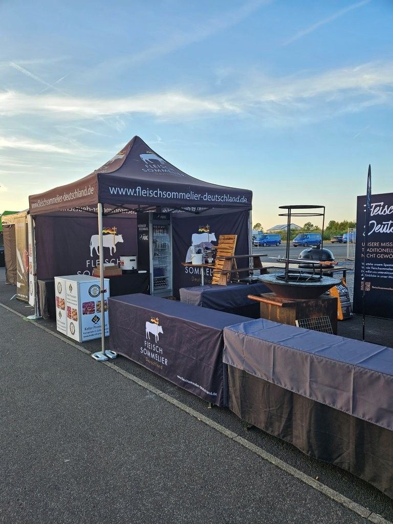 Ein Grillstand im Freien mit bordeauxroten Zelten und Tischen, die bei Sonnenuntergang auf einem gepflasterten Platz aufgebaut sind.