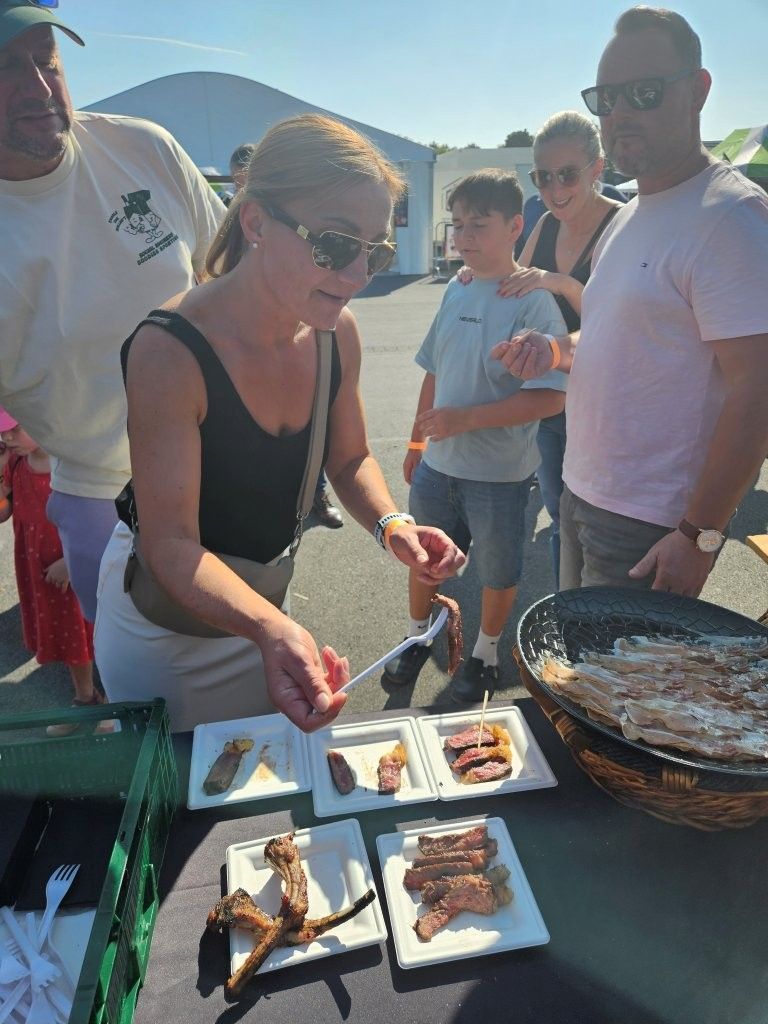 Eine Frau serviert Grillgerichte von einem Tisch bei einer Feier im Freien, während Menschen in der Nähe im hellen Sonnenlicht stehen.