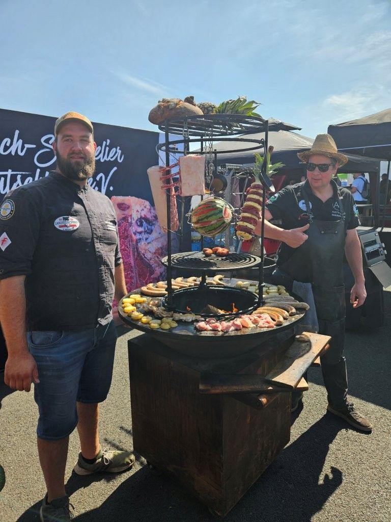 Zwei Personen stehen neben einer großen Grillanlage im Freien bei einer sonnigen Veranstaltung, auf der Speisen auf Spießen gegrillt werden.